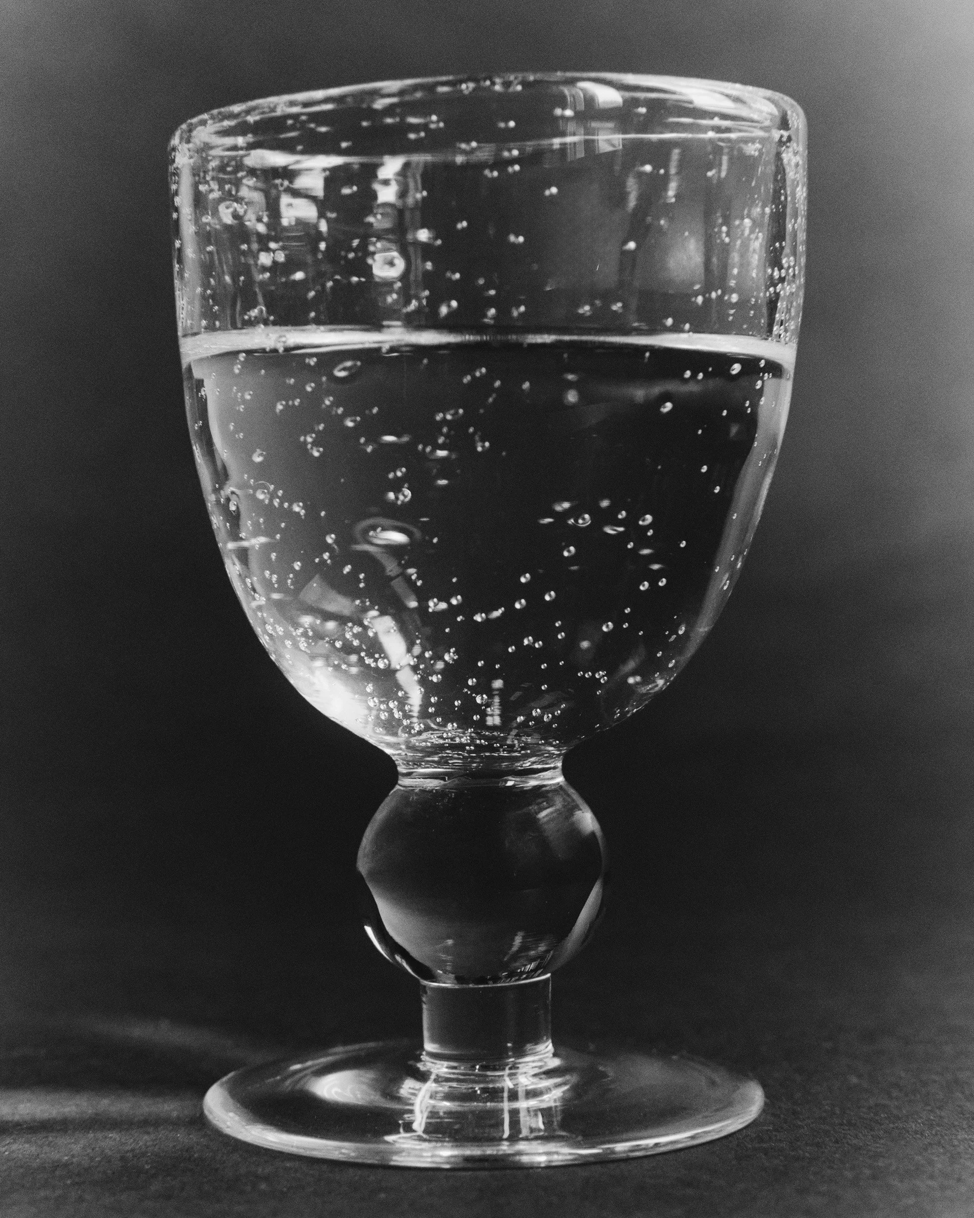 A close-up black-and-white photo of a glass of sparkling water with bubbles, placed on a dark surface against a dark background.