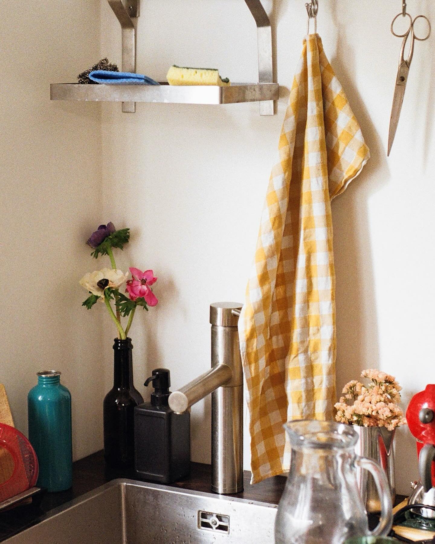 A kitchen corner with a stainless steel shelf holding a sponge and scrubbing pad, yellow and white checkered dish towel hanging from a hook, a black soap soap dispenser, a blue bottle, and a small black container near the sink, which has a silver fau