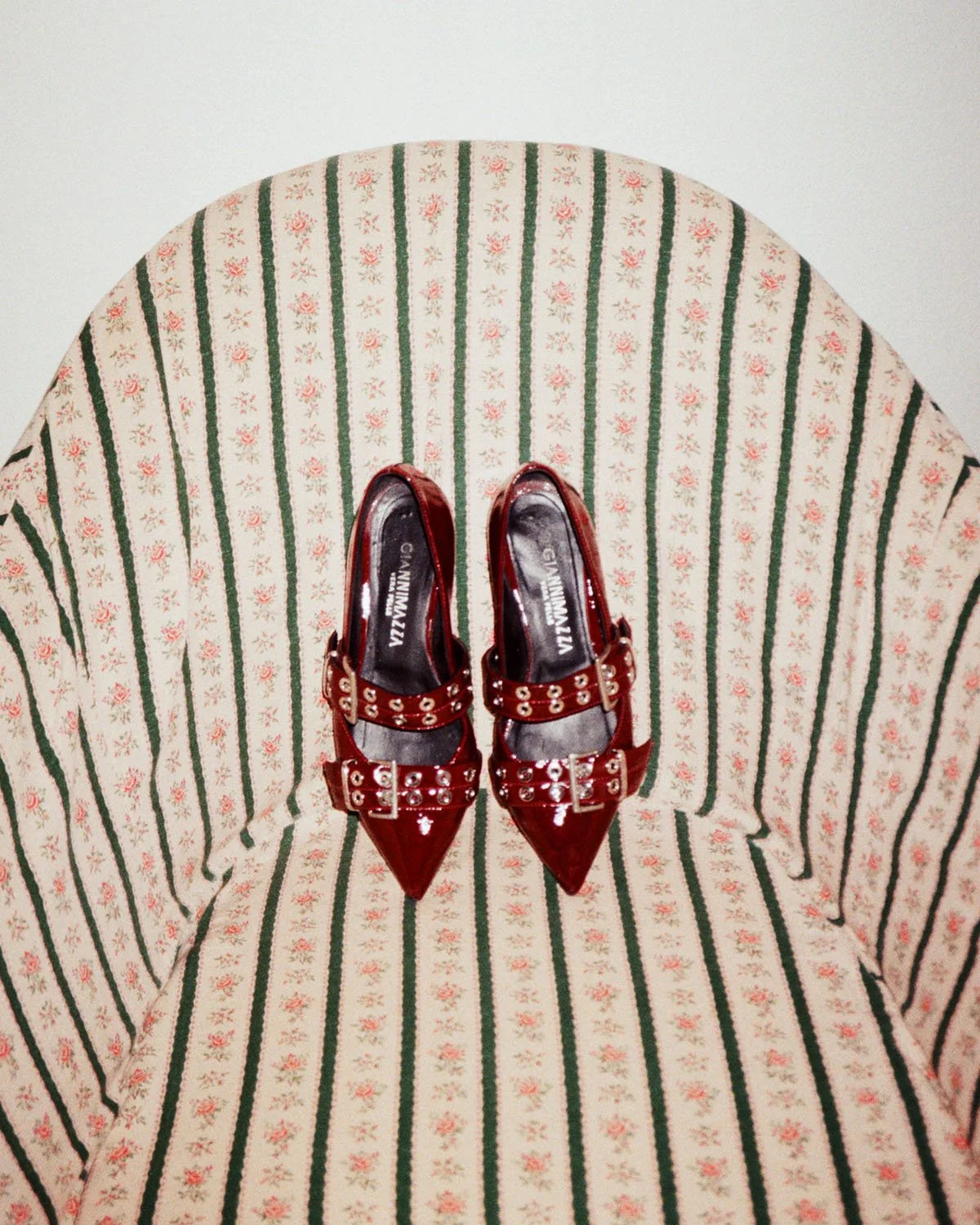 Red patent leather high-heeled shoes with silver embellishments on a green and red striped upholstered chair.
