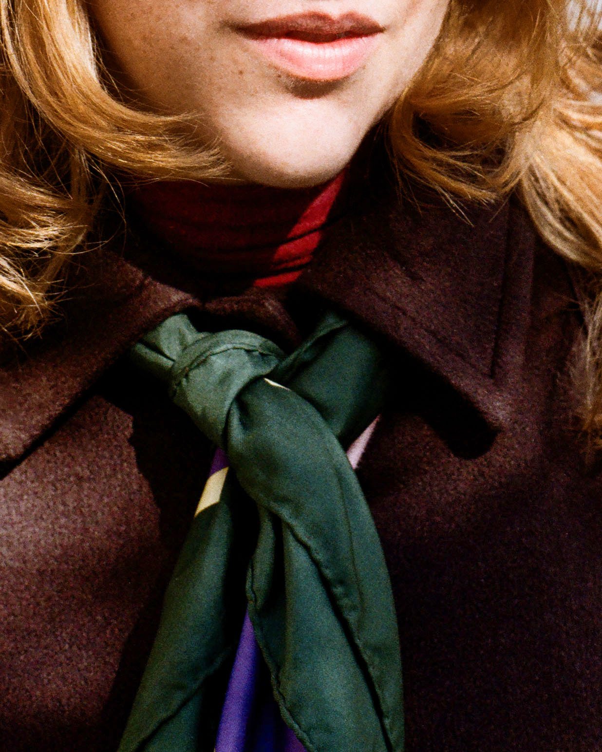 Close-up of a person's face, focusing on lips and chin, with red hair, wearing a dark brown coat, red shirt, and a green scarf tied around the neck.