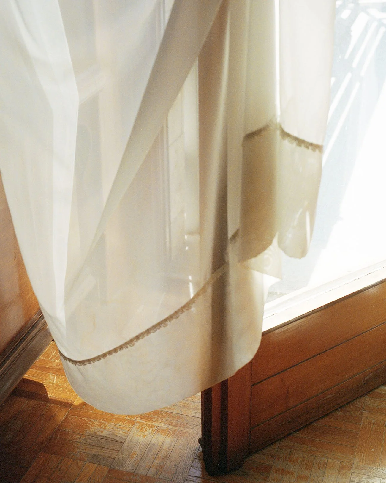 View of a beige curtain hanging in front of a window with light coming through, with wooden furniture and flooring visible.
