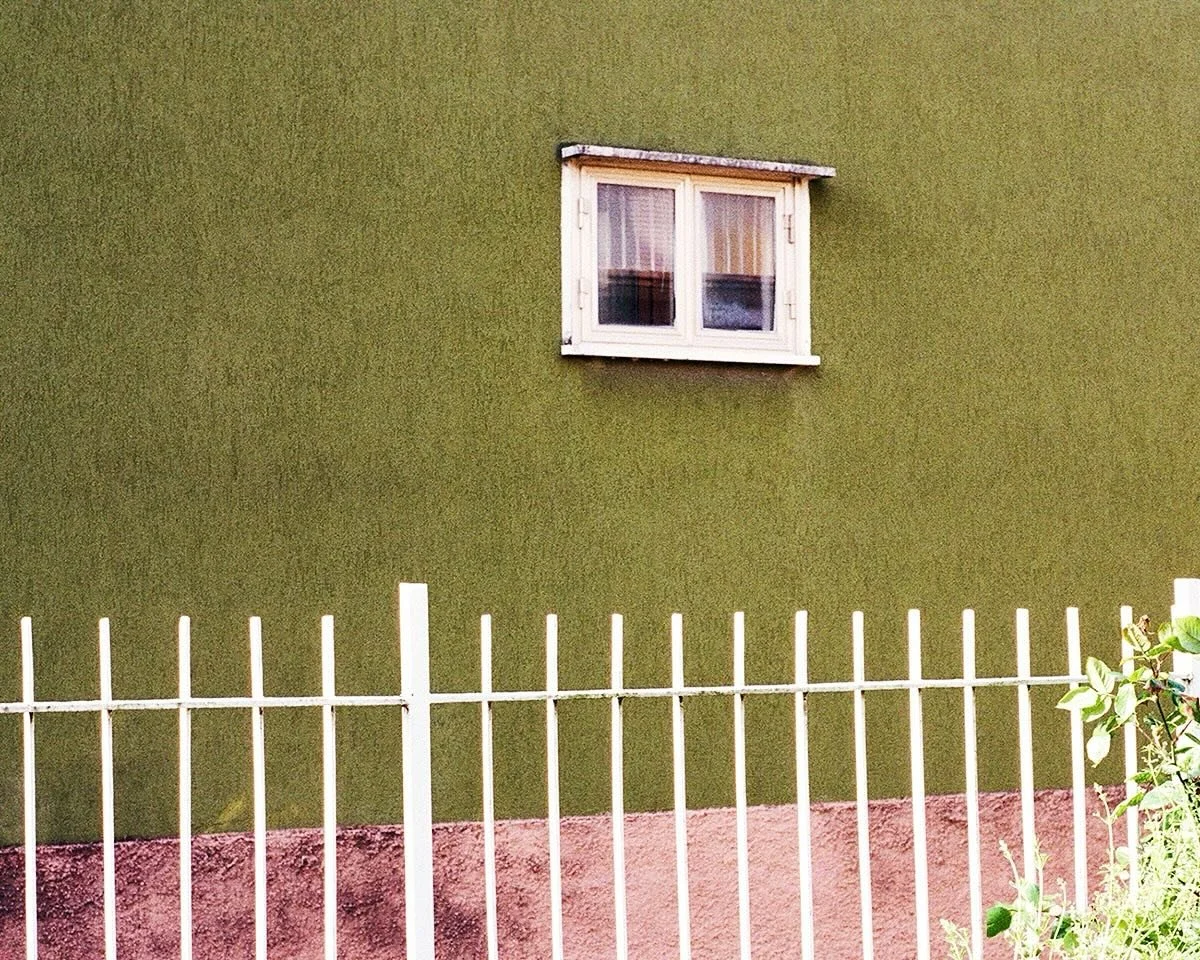 Green exterior wall with a small white-framed window and a white picket fence in front.