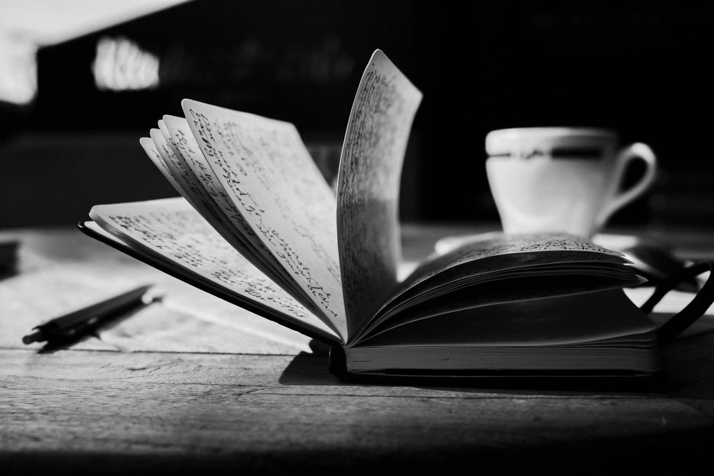 Open notebook with handwritten text on pages, set on a wooden surface, with a pen lying next to it, and a coffee cup in the background, all in black and white.