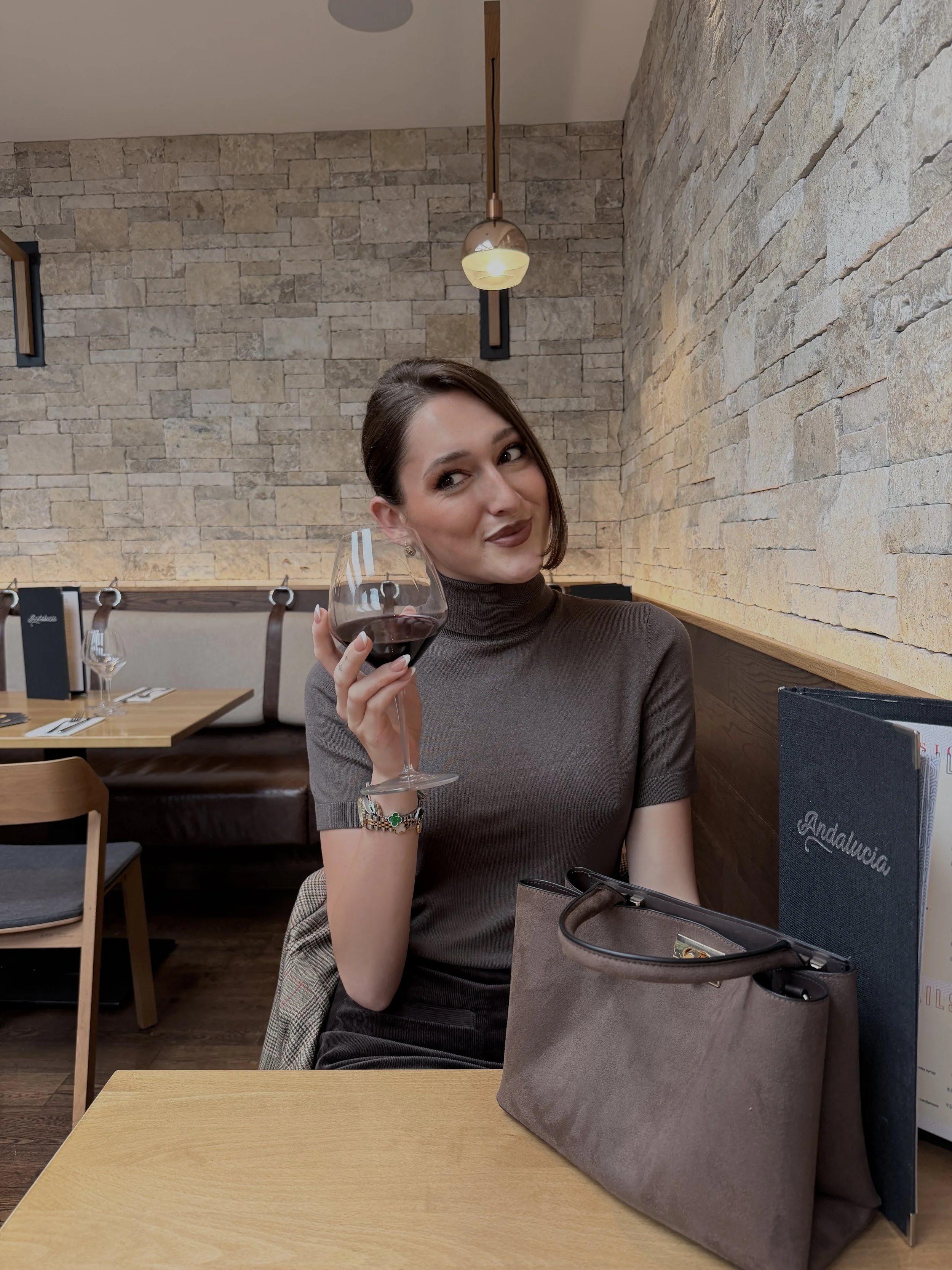 A woman sitting at a restaurant table holding a glass of red wine, looking to her left with a playful smirk, with a beige wall and modern light fixture behind her, a menu and a purse on the table.