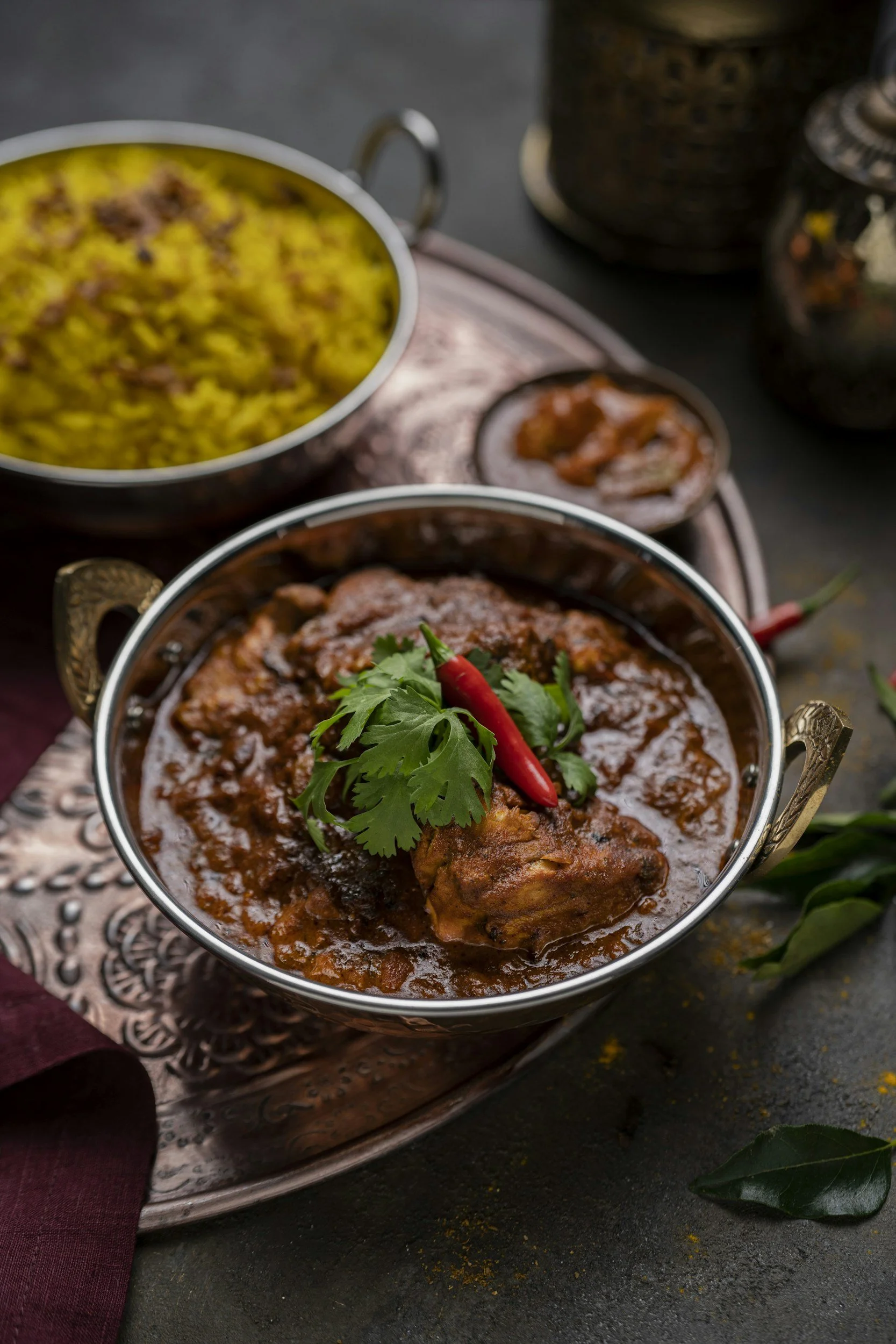 A bowl of Indian chicken curry garnished with cilantro and a red chili, with a side of yellow rice and other spices in the background.