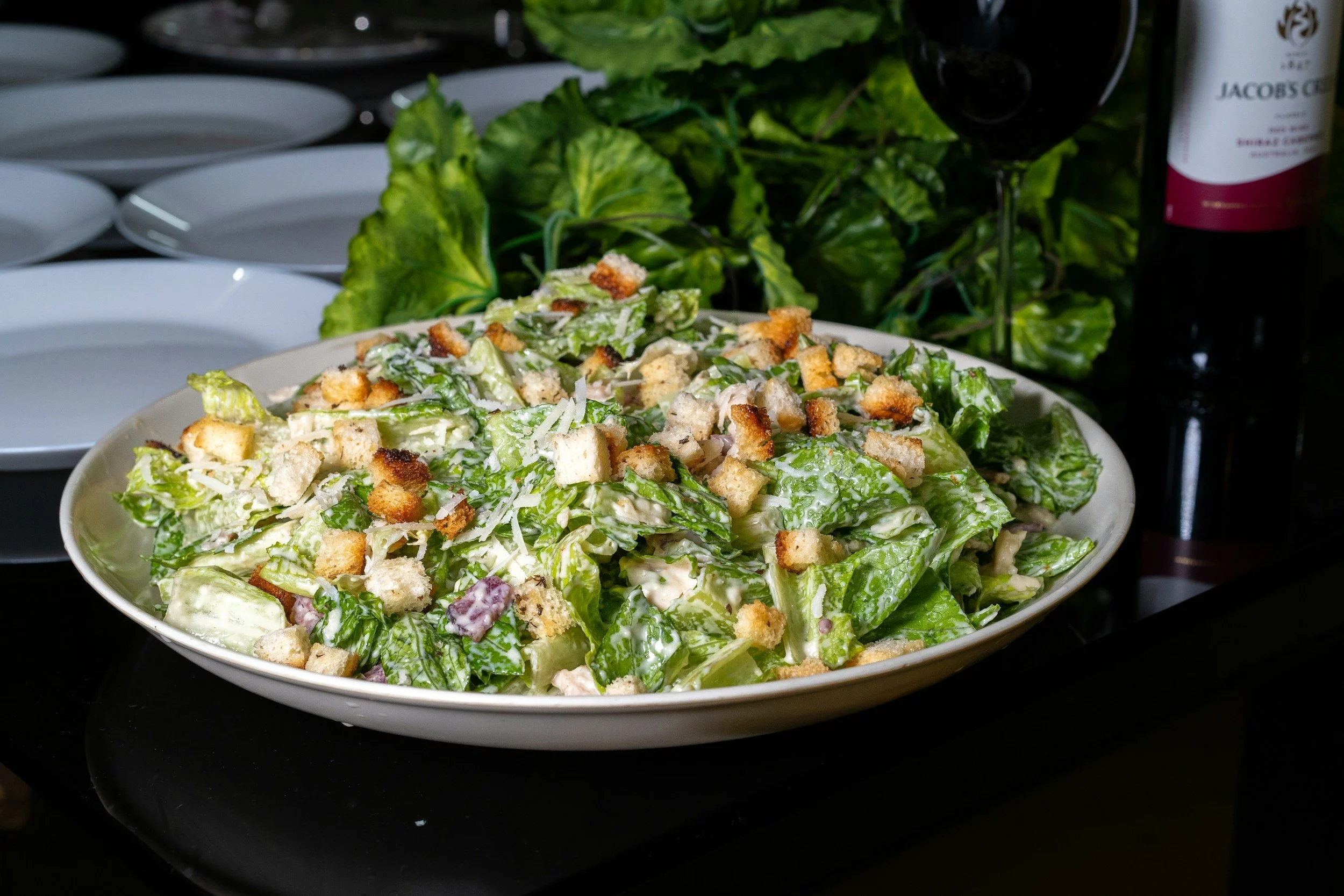 A white bowl of Caesar salad with chopped romaine lettuce, grated cheese, croutons, and dressing, placed on a black surface.