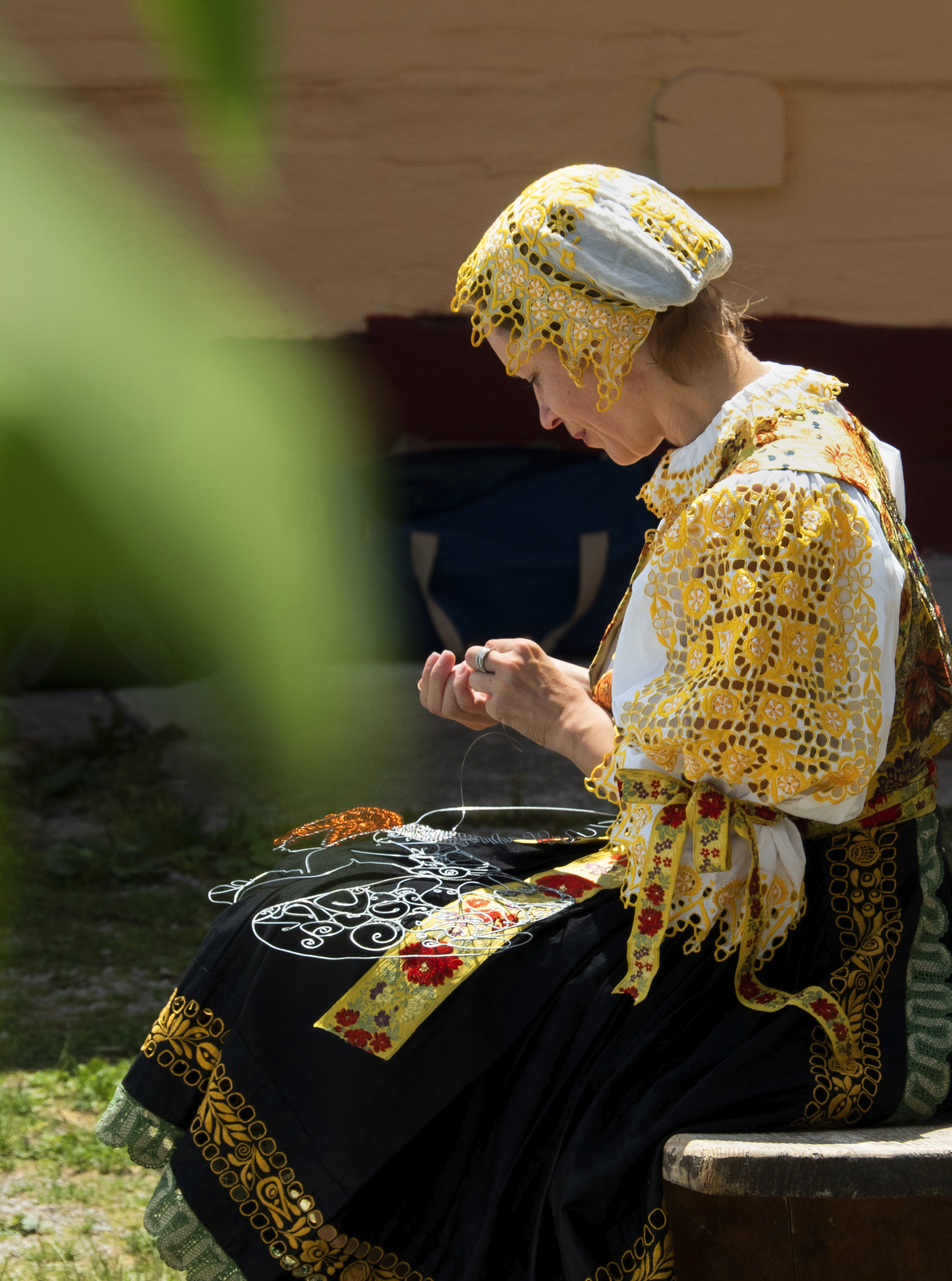 Woman wearing a yellow embroidered dress and a matching headscarf, sitting outdoors with embroidered jewelry and textiles displayed in front of her.