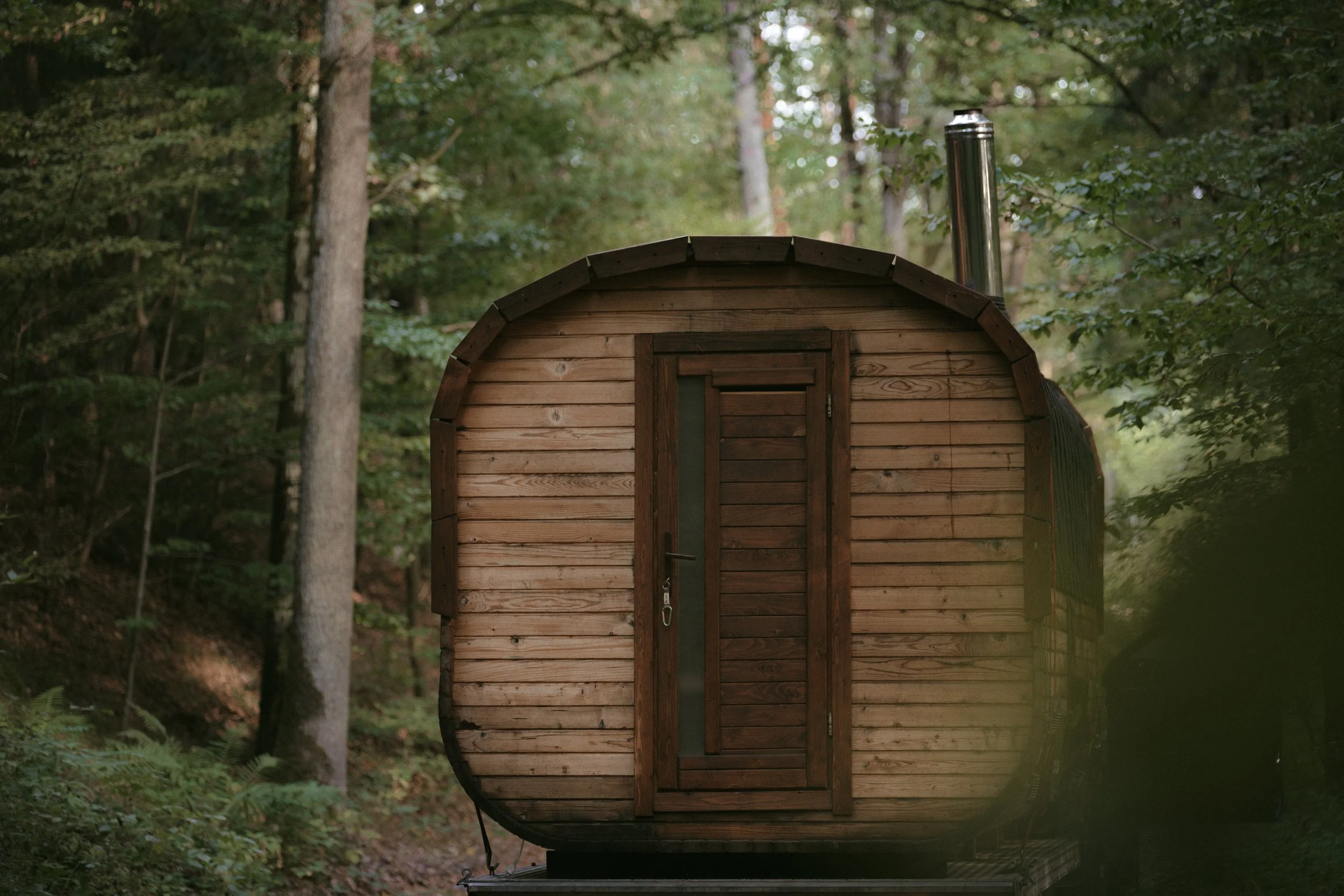 Ein kleines, holzverkleideter Saunafass in einem grünen Wald im Hintergrund.