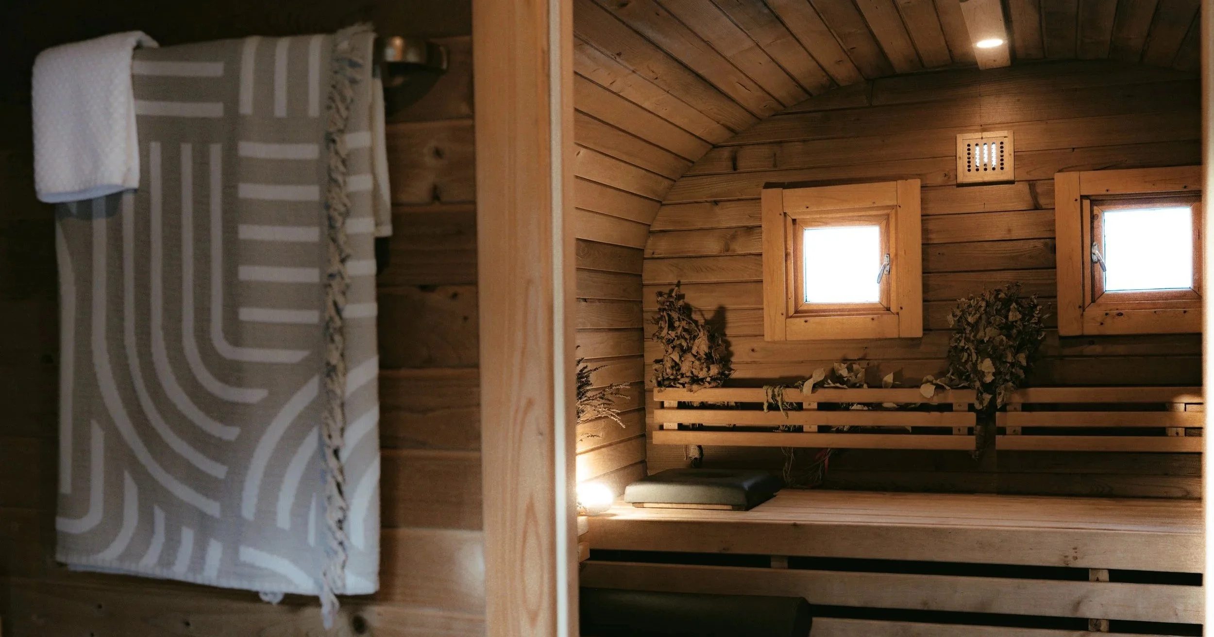 Ein Saunaraum mit Holzvertäfelung, zwei kleinen Fenstern und einer Holzbank, dekoriert mit trockenen Pflanzen und Steinen.