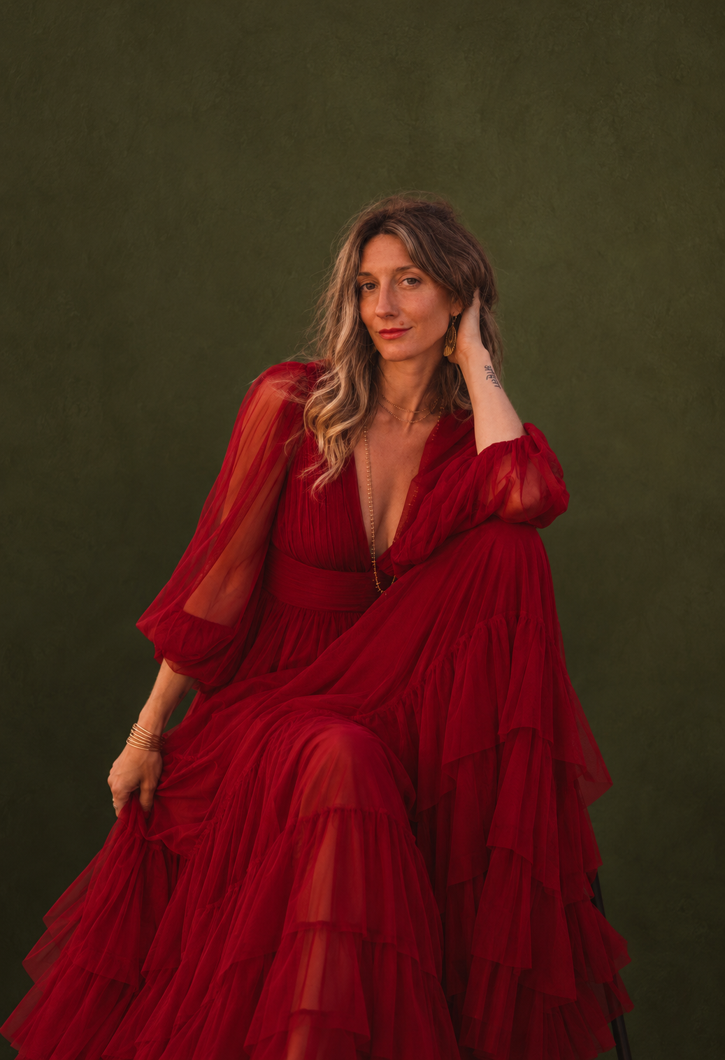 A woman in a long, flowing red dress sitting against a dark green background, with one hand resting on her knee and the other touching her hair.