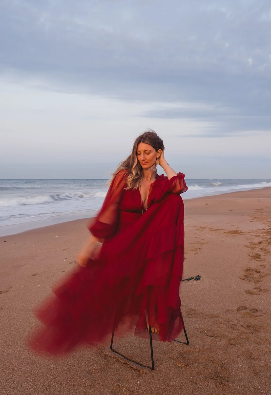 Woman in a long red dress sitting on a chair on the sandy beach with ocean waves and cloudy sky in the background.