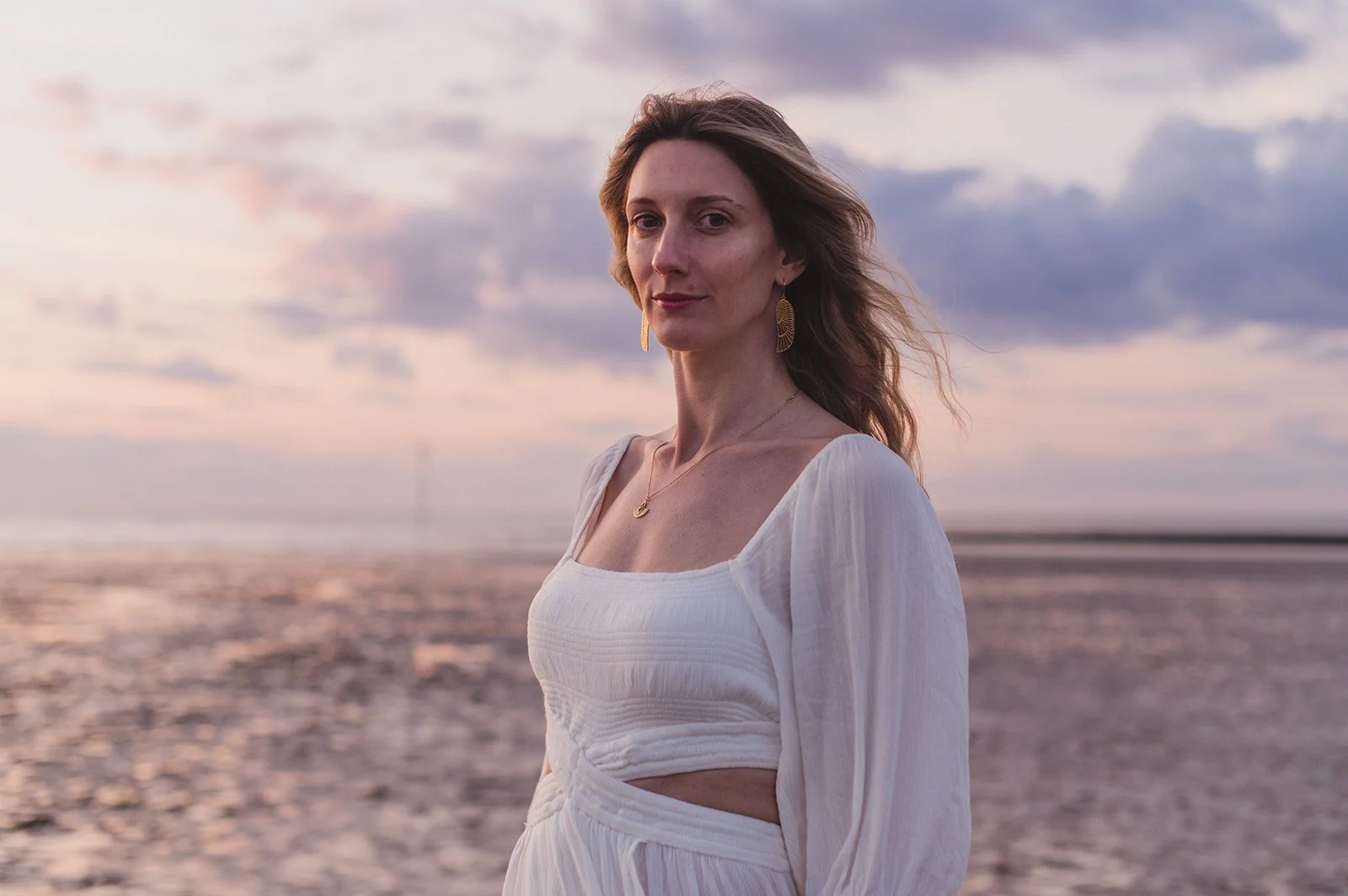 A woman with long, wavy brown hair wearing a white dress with cutouts, standing on a beach during sunset. She has gold earrings and a delicate necklace, with a calm expression on her face.