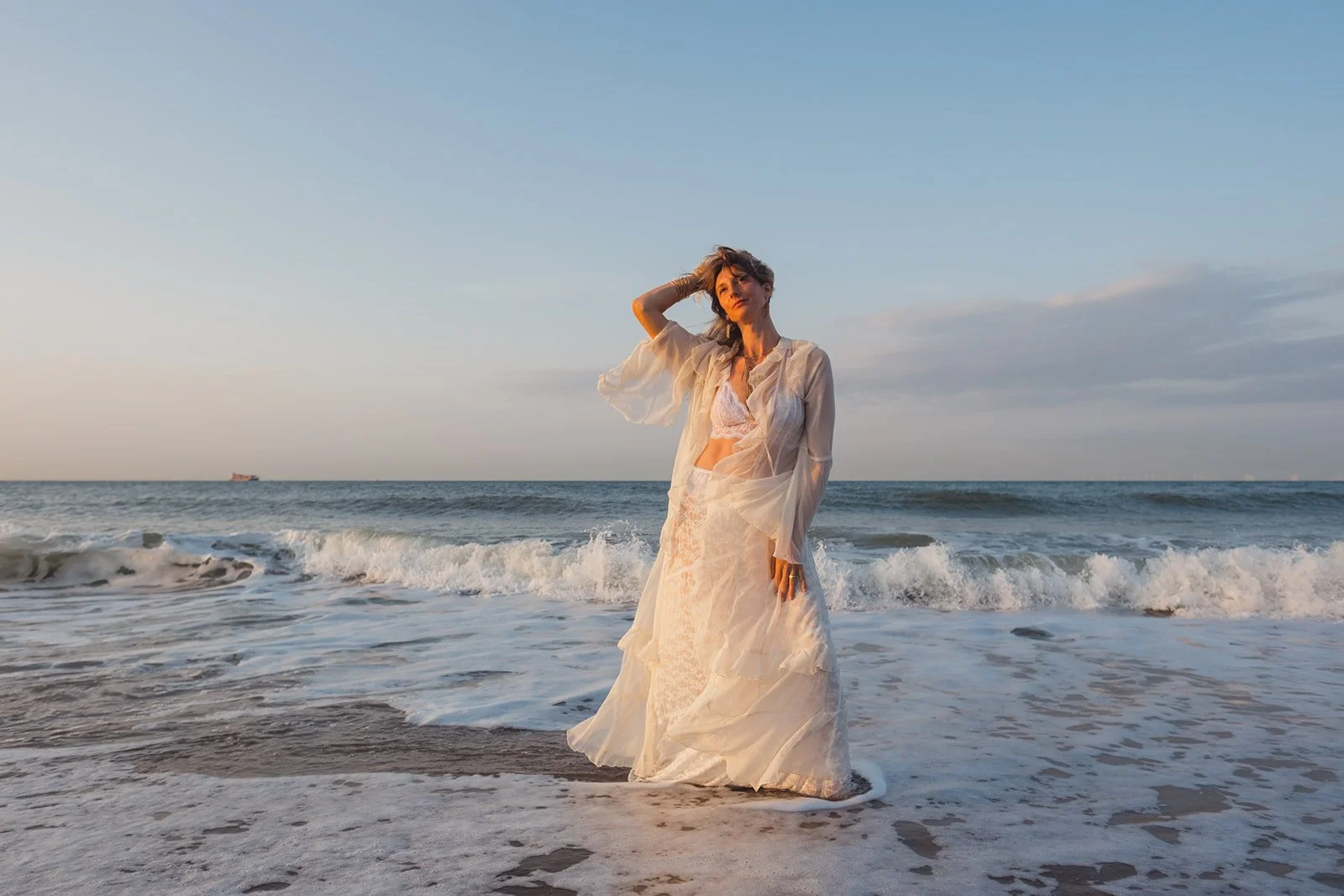 Woman in a white dress standing in the ocean waves at sunset.
