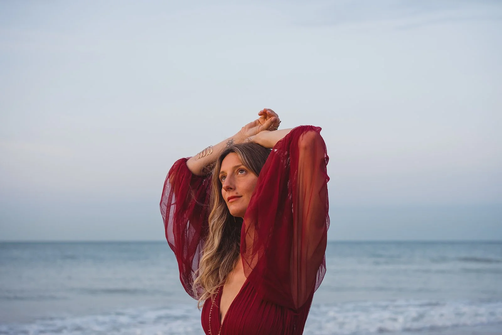 Woman in red dress standing on the beach with arms raised above head, gazing into distance, ocean in background, overcast sky.
