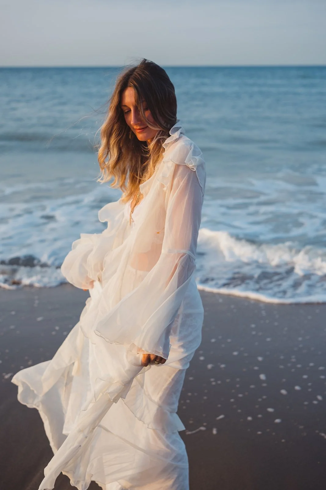 A woman wearing a sheer white dress stands on the beach near the ocean waves during sunset.