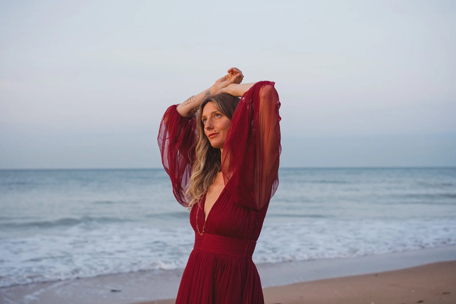 A woman in a red dress with sheer sleeves stands on a beach with her arms raised and hands clasped above her head, looking to the side, with the ocean and an overcast sky in the background.