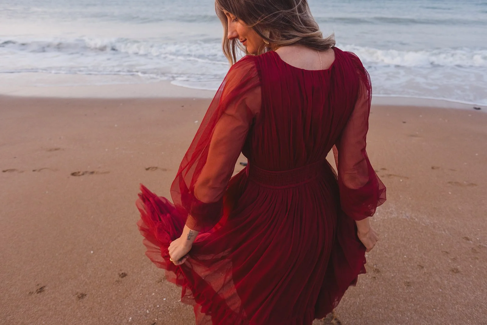 Woman in a flowing red dress walking on the beach near the ocean with waves in the background.