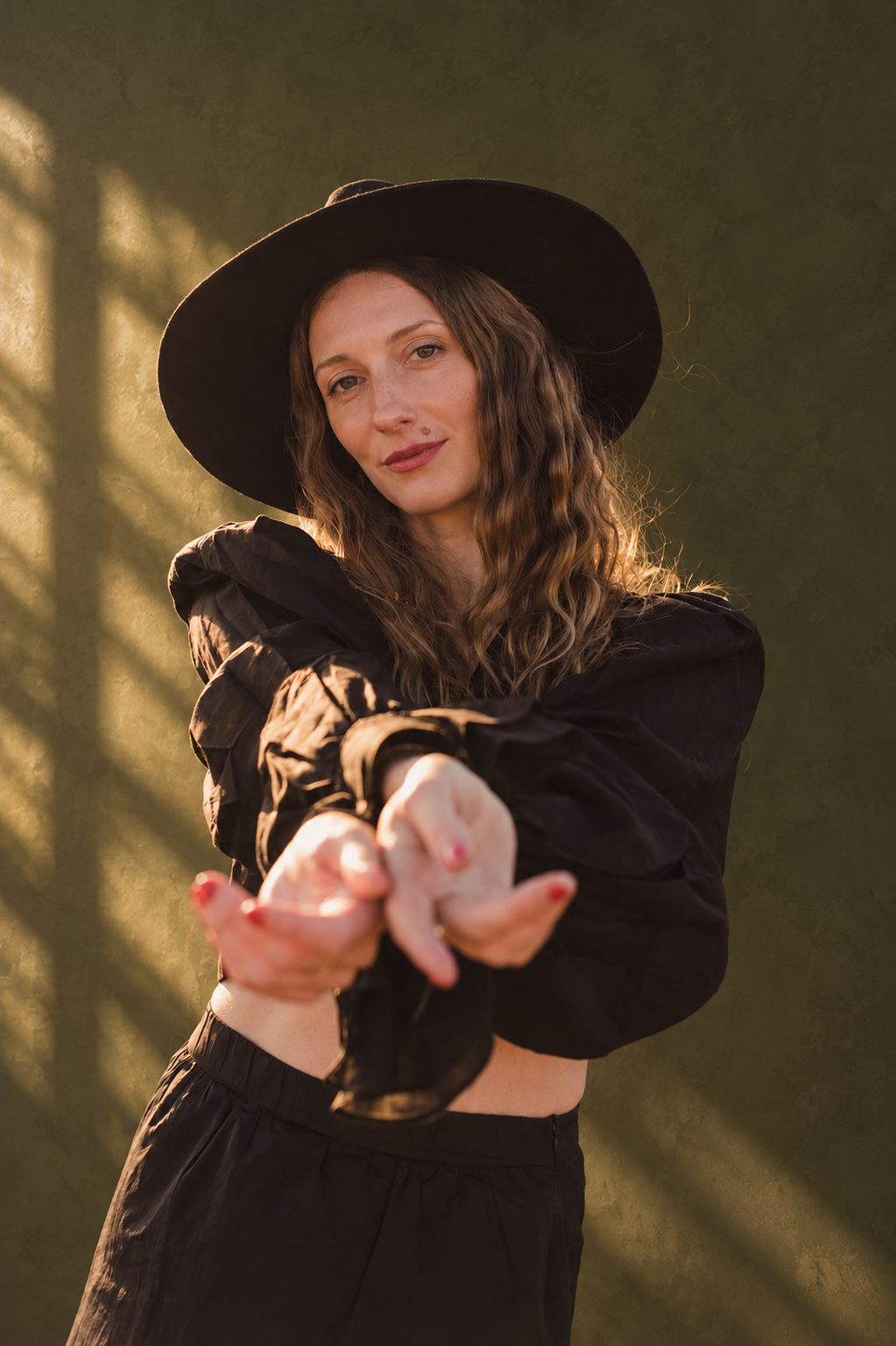 A woman with long, curly hair wearing a black wide-brimmed hat and black outfit, pointing or reaching toward the camera, standing against a textured green background with warm lighting.