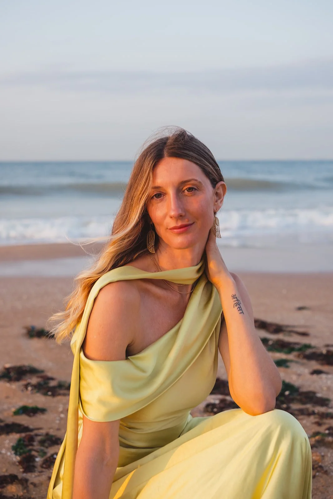 A woman in a yellow dress sitting on the beach during sunset, with the ocean and sky in the background.