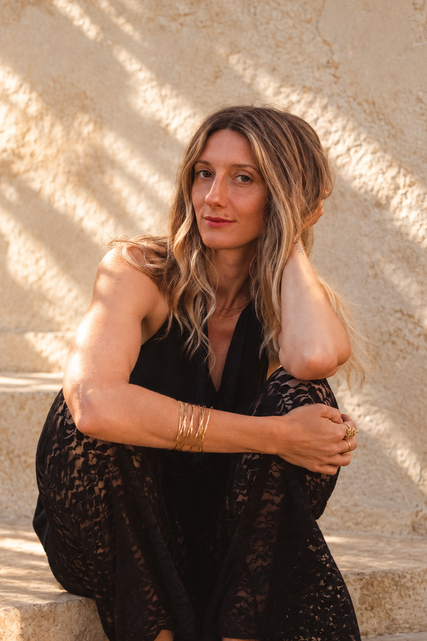 A woman with wavy blonde hair, sitting on stone steps in sunlight, wearing a black sleeveless dress with lace, gold jewelry, and looking into the camera with a relaxed expression.