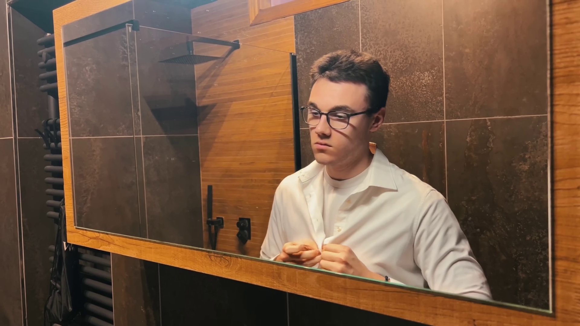 Seba si abbottona una camicia bianca e si guarda allo specchio nel bagno di casa sua. Immagine di anteprima tratta dal cortometraggio "Caesena me cecidit".