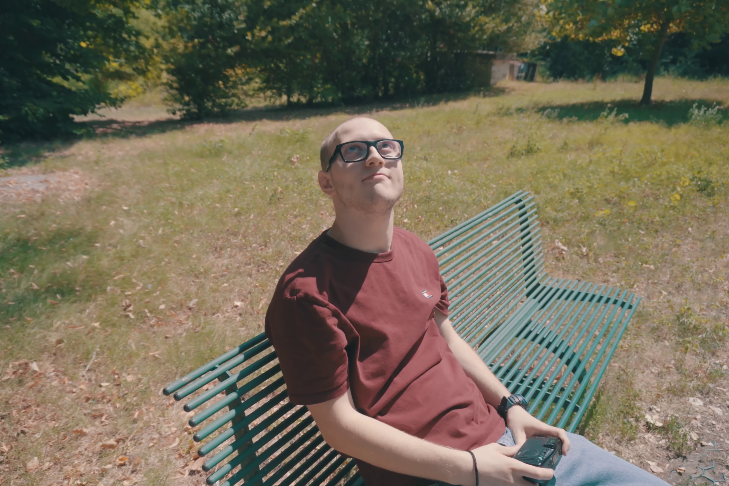 Stills sta seduto su una panchina in un parco sotto alberi verdi in estate, vestito con una maglia color rosso scuro e jeans blu. Immagine di anteprima tratta dal cortometraggio "Black, White & Stills".