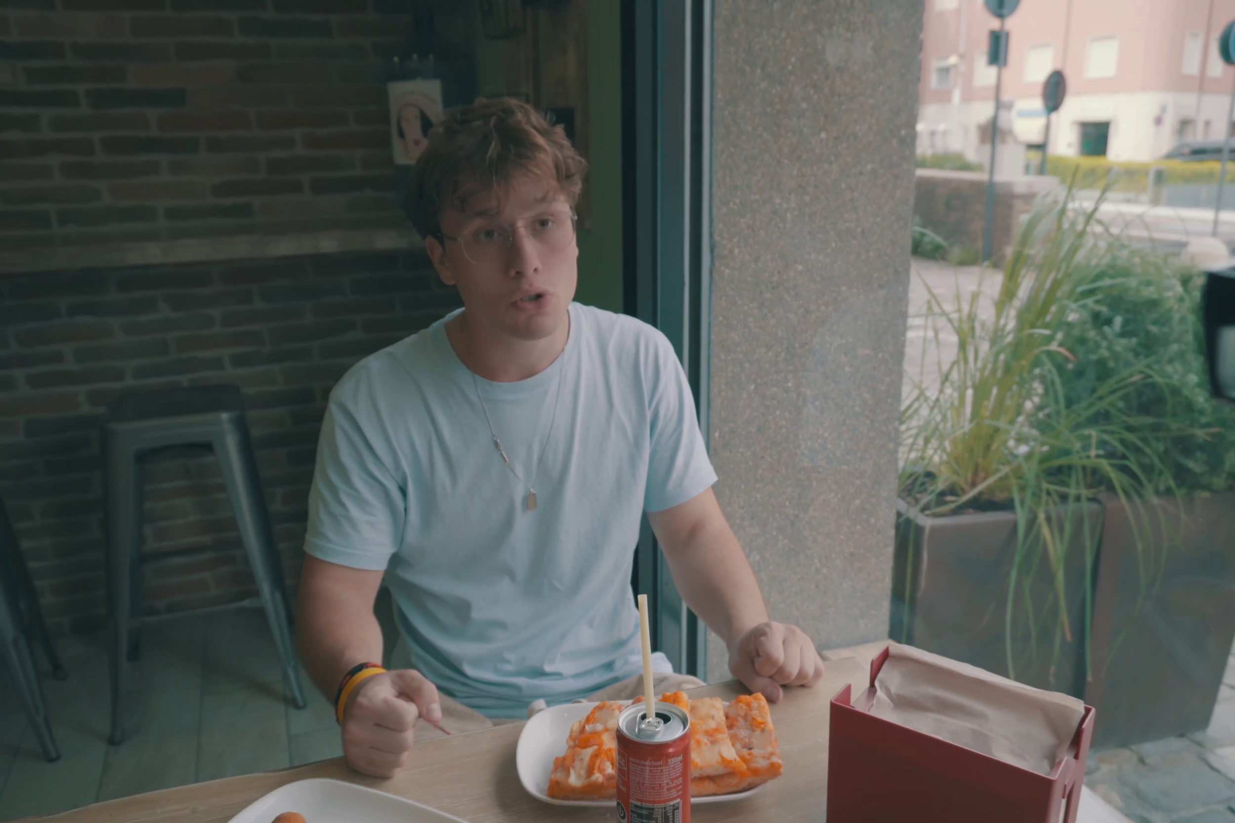 Max parla a Stills in una inquadratura che fa vedere anche la città oltre la vetrata della pizzeria, Max è vestito con una maglietta azzurra, come la parete della pizzeria. Immagine di anteprima tratta dal cortometraggio "Black, White & Stills".