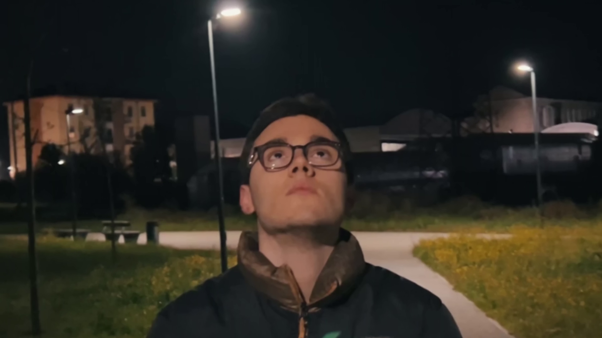 Seba pedalando su una bicicletta guarda verso l'alto di notte in un parco, con lampioni e case sullo sfondo. Immagine di anteprima tratta dal cortometraggio "Caesena me cecidit".