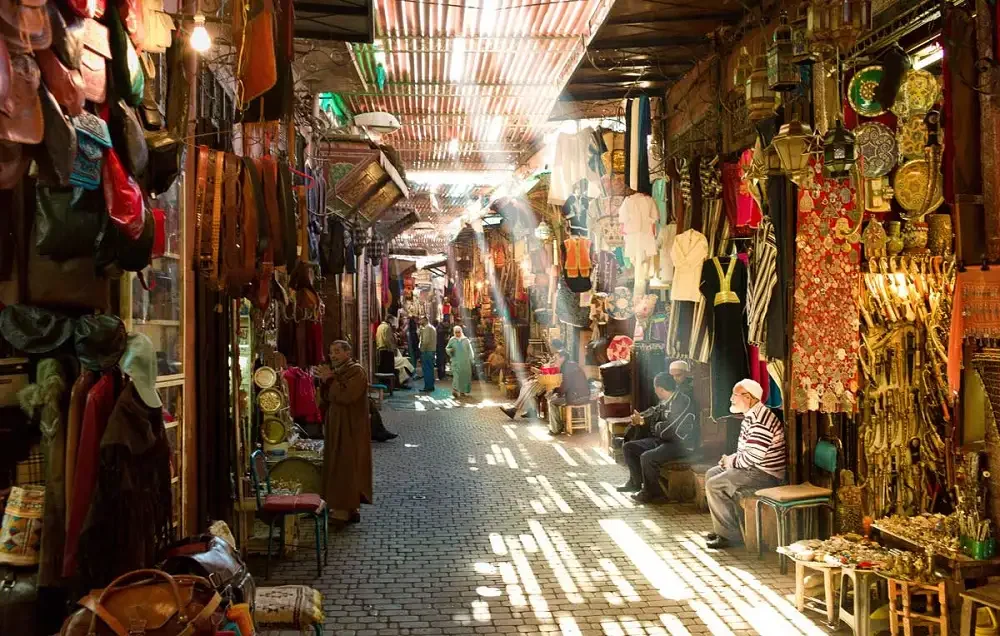 A lively marketplace alley with stalls on both sides selling clothing, bags, and decorative items. Shoppers and vendors sit and browse, with sunlight filtering through the narrow aisle.