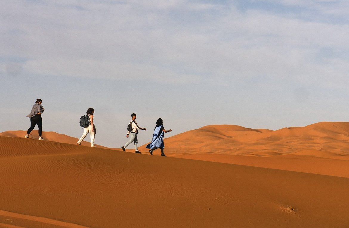 Five people walking in a line across sand dunes in a desert under a cloudy sky