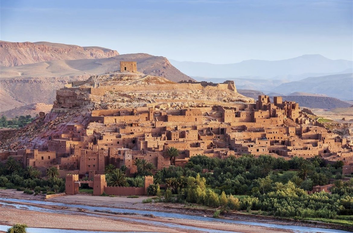 Ancient kasbah or fortress built on a hillside with traditional mud-brick buildings, surrounded by green oasis and mountains in the background