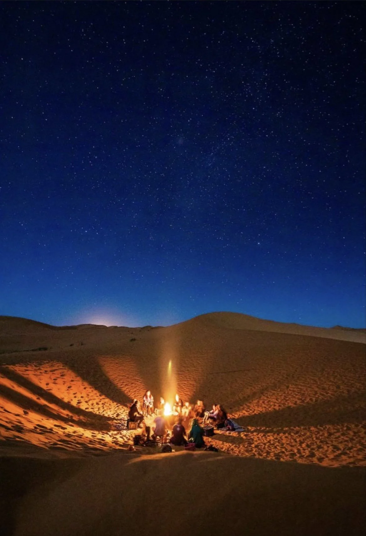 A group of people sitting around a campfire in a desert at night, with a dark sky filled with stars above.