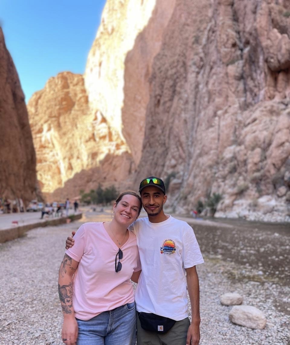 A smiling woman and man standing together in a rocky canyon, with tall steep cliffs on either side, in a desert landscape during daylight.