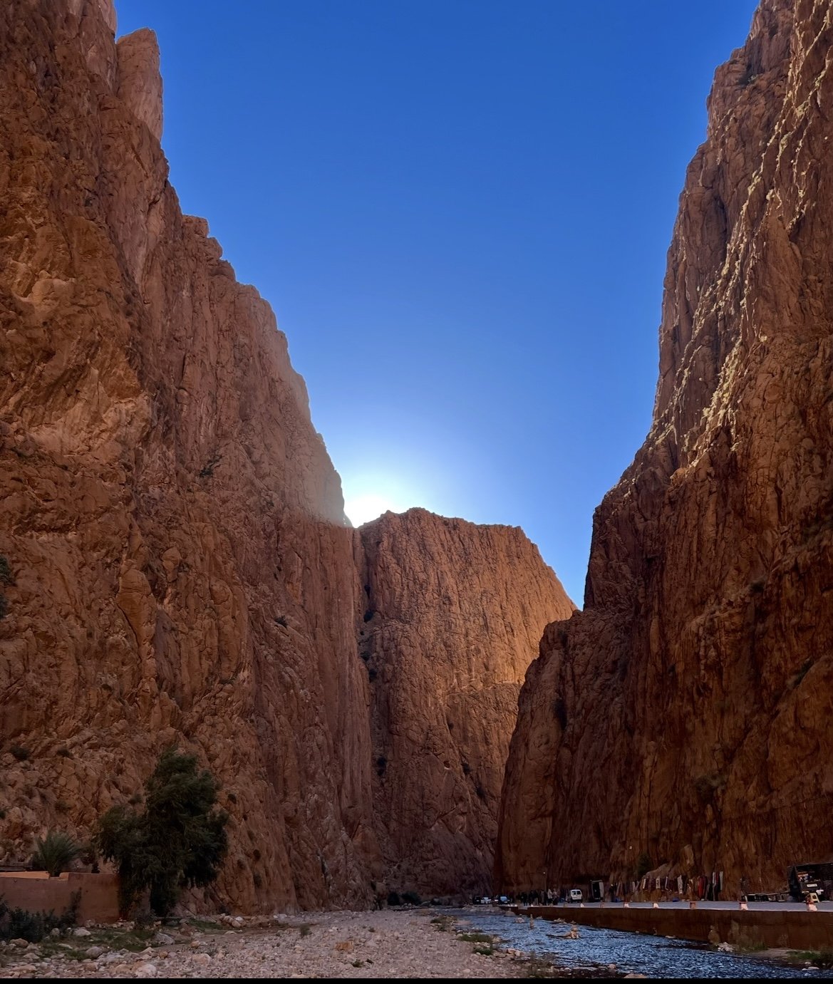 The image shows a deep canyon with tall, reddish-brown rock walls on either side and a narrow river flowing through the bottom. The sky above is clear and blue, with the sun partly hidden behind the rocks.