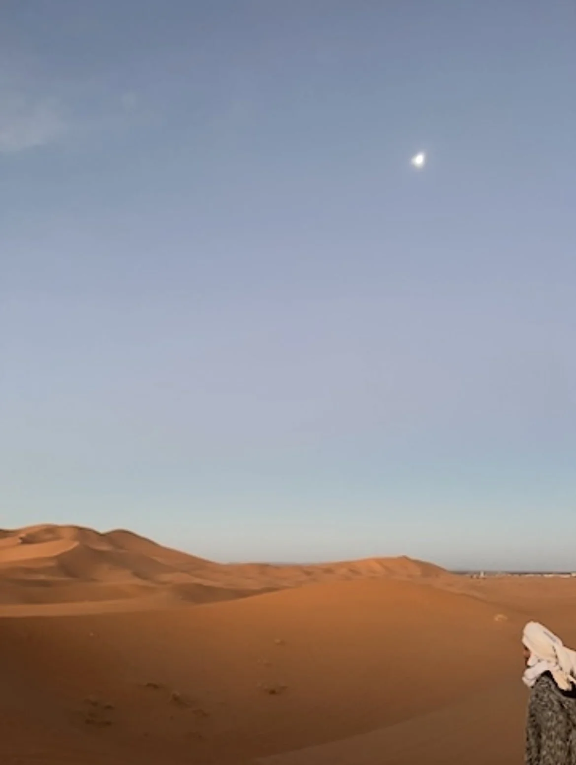 Desert landscape with sand dunes under a clear sky and a small moon or planet visible in the upper right portion of the sky, with a person standing in the lower right corner.