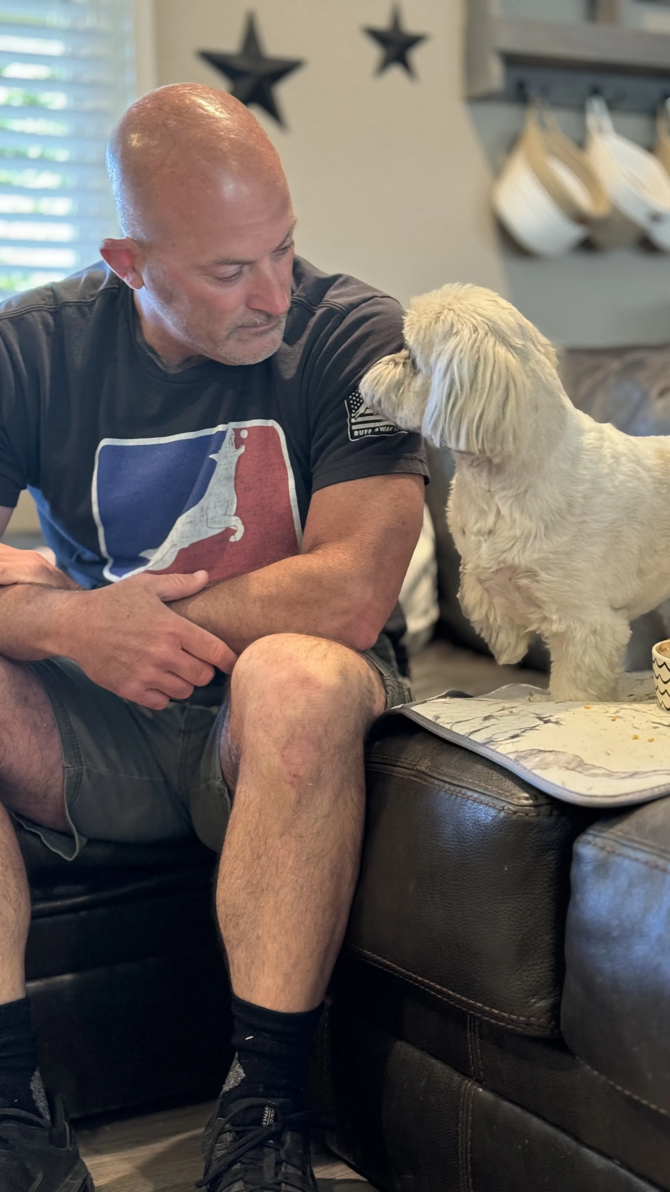 A bald man in a black T-shirt with a Texas-themed graphic is sitting on a brown leather couch. He is looking down at a light-colored puppy that is leaning on his arm. The background includes a window with blinds, decorative stars on the wall, and hanging white items.