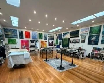 An art gallery with landscape paintings on the walls, a table with a white tablecloth, and a barrier with stanchions in the center of the room, wooden flooring, and skylights on the ceiling.