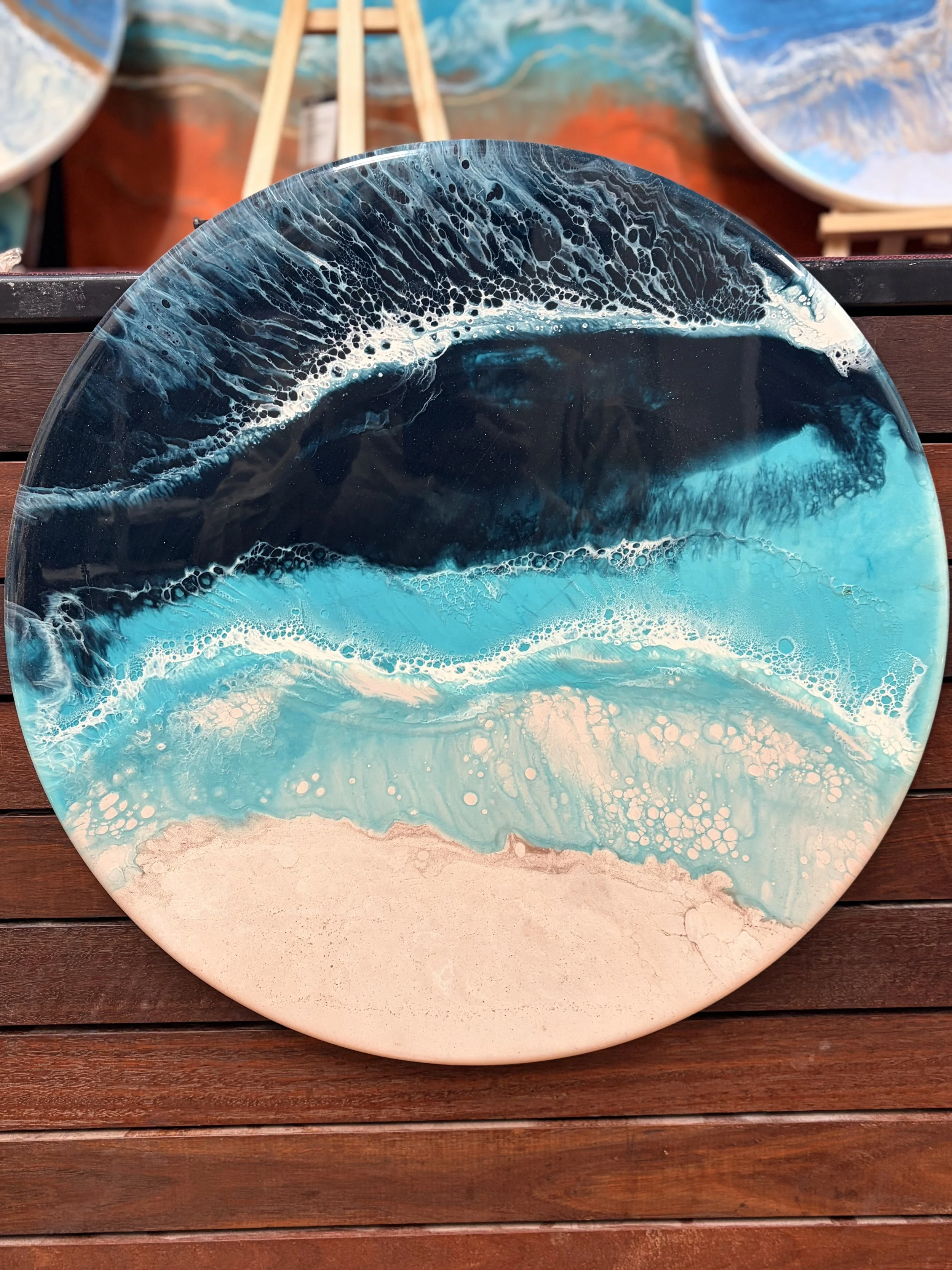 Round decorative tray with ocean wave pattern in shades of blue, white, and turquoise, placed on a wooden surface with other similar trays in the background.