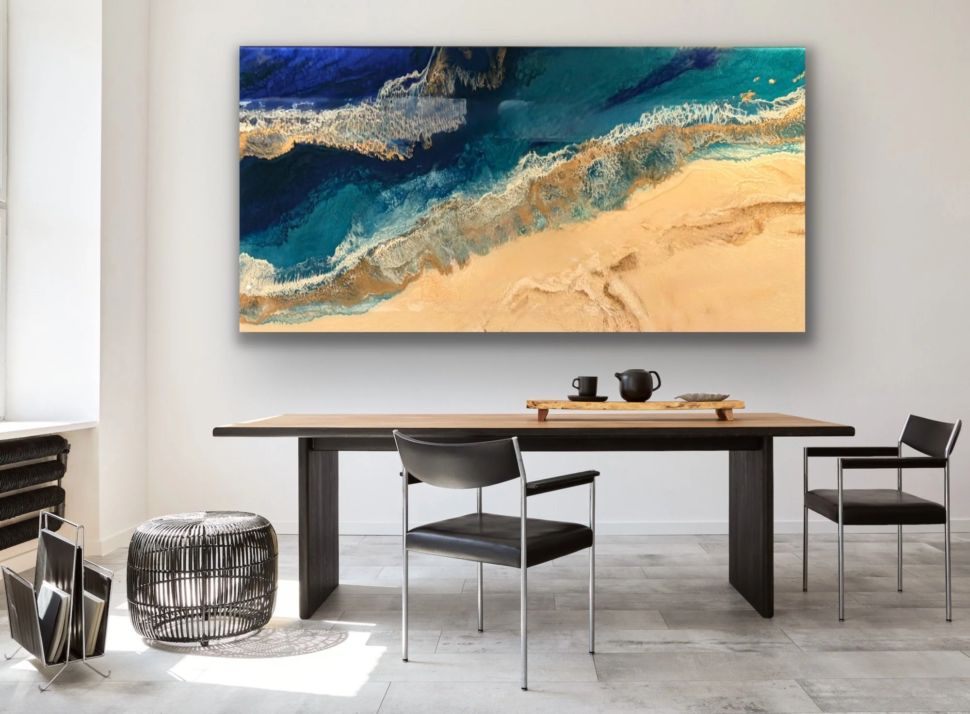 Interior of a modern dining area with a black table, black and gray chairs, a wooden tray with black teaware, a black rattan basket, and a large abstract ocean-themed painting on a white wall.