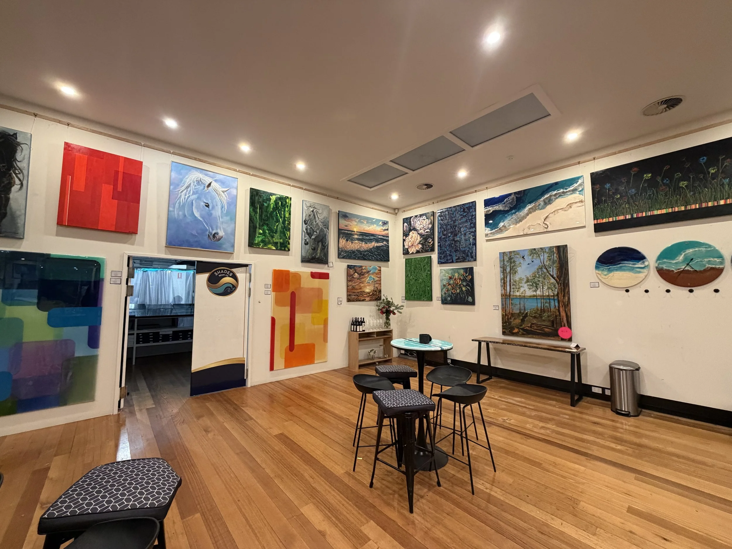 Interior of an art gallery with various paintings on white walls, a few small tables with chairs, wood flooring, and overhead lighting.