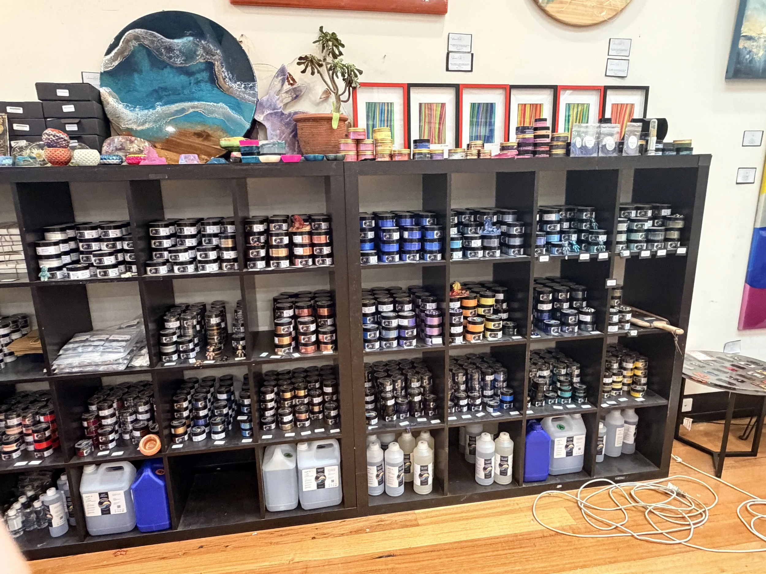 Black shelving unit displaying various art supplies such as jars of colored powders or paints, bottles of paint or cleaning solutions, and stacked small containers. Decorative artwork and plants are on top of the shelf.
