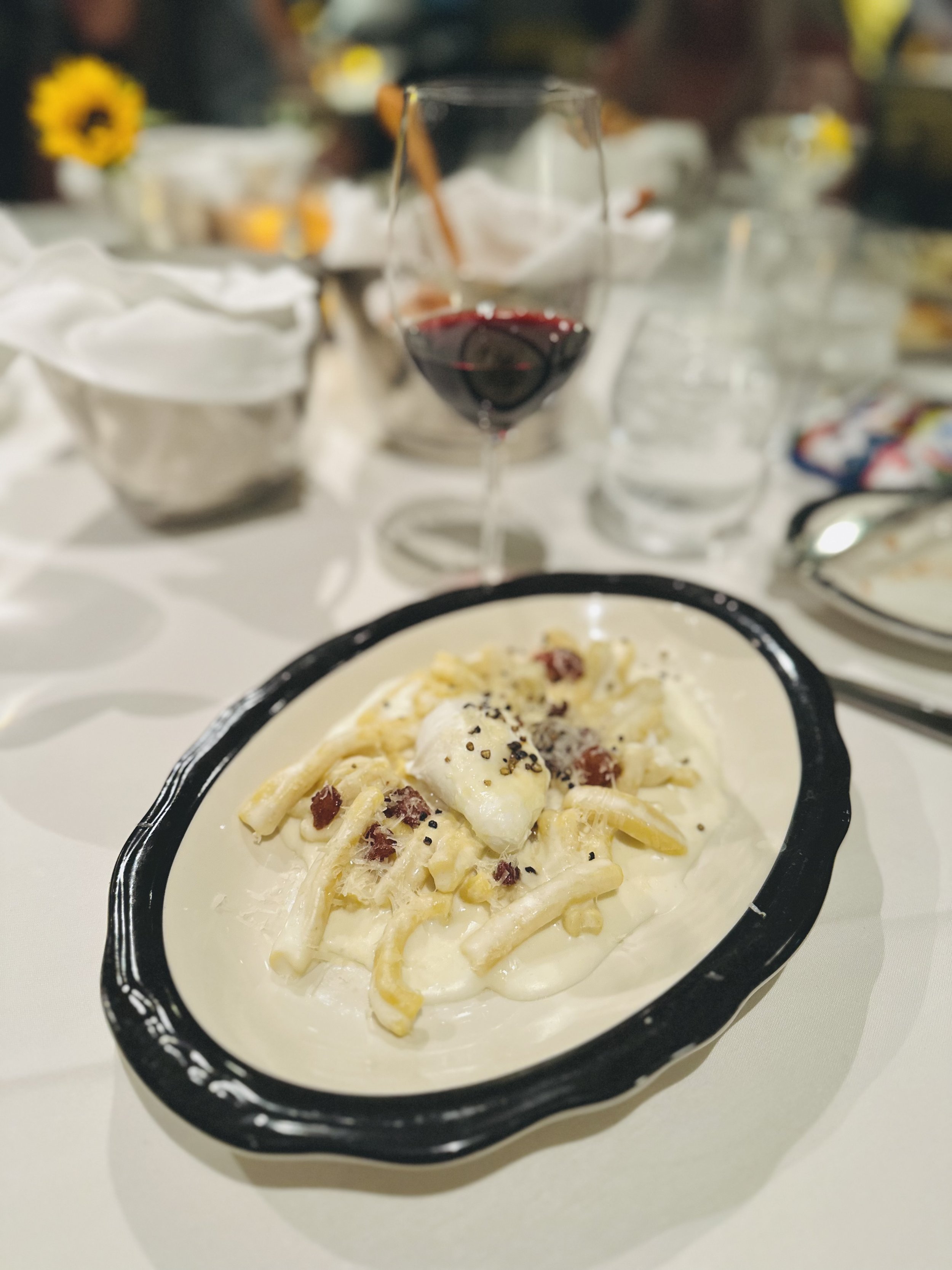 Bowl of creamy pasta with bacon bits, topped with black pepper, on a white table. In the background, a glass of red wine and other dishes are visible.