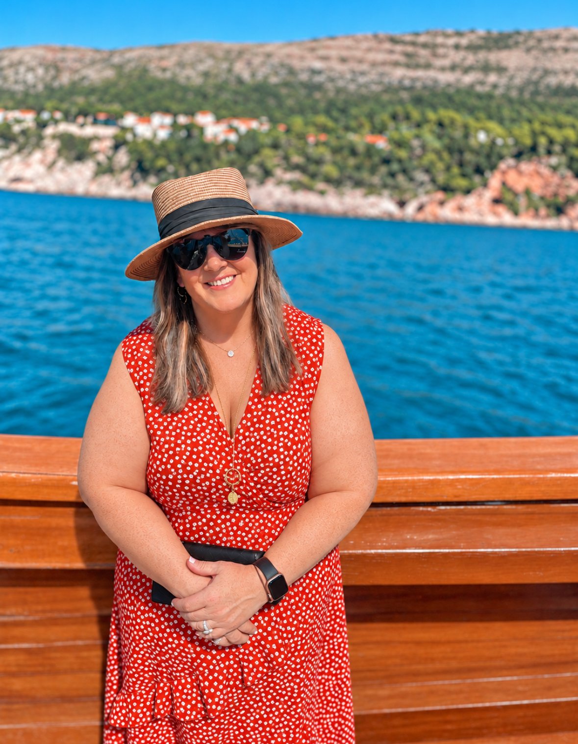 Woman wearing a red polka dot dress, straw hat with black ribbon, and sunglasses, smiling on a boat with blue water and hilly landscape with houses in the background.