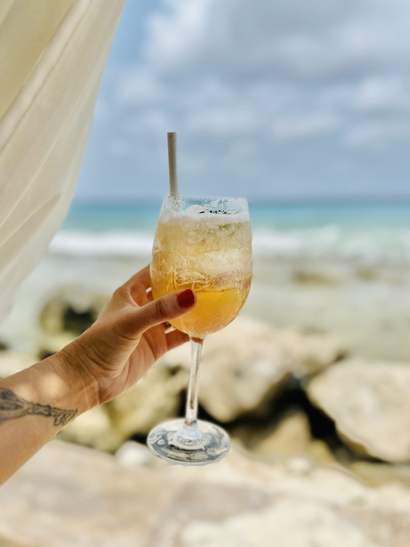 A person holding a glass of a cold, golden beverage with ice and a straw, at the beach with a cloudy sky in the background.
