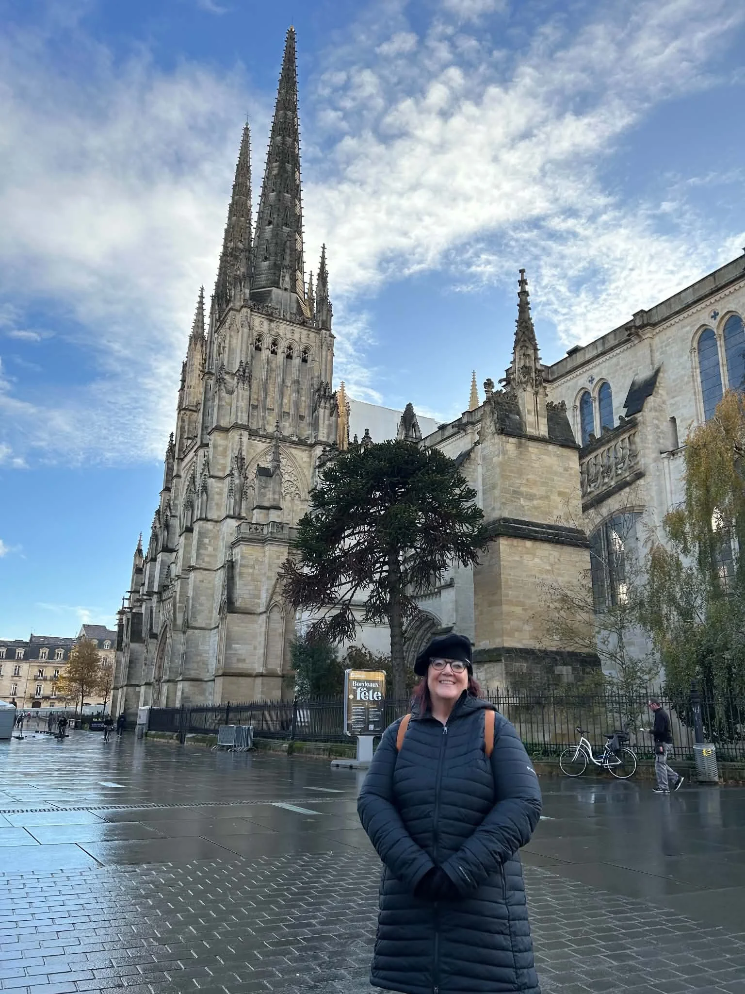 A woman with glasses and a black coat standing on a wet cobblestone street in front of a large Gothic cathedral with tall spires. The sky is cloudy with some patches of blue.