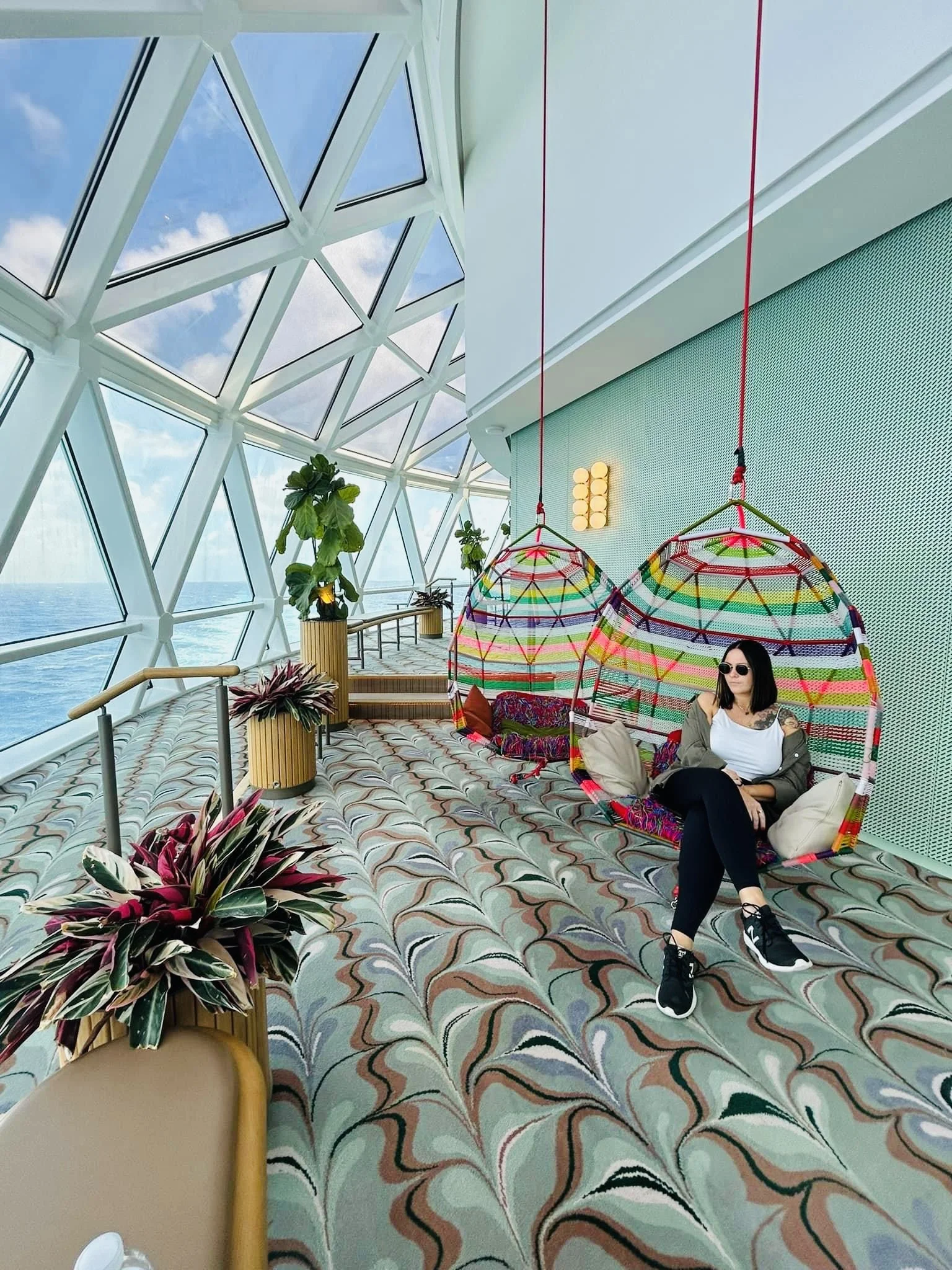 A woman sitting on a colorful hanging chair inside a modern glass building with ocean views, plants, and patterned carpet.
