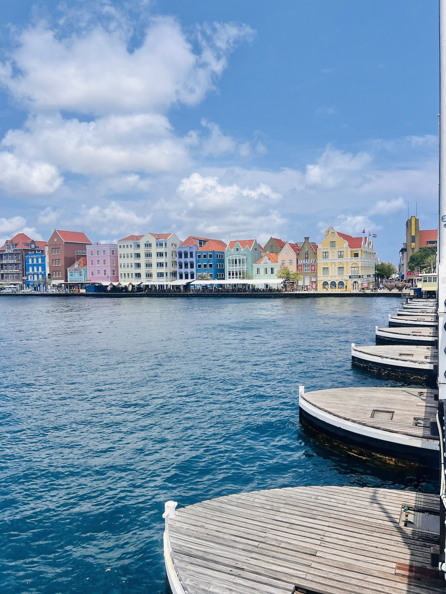 Colorful row of European-style buildings along a waterfront, with a wooden dock and water in the foreground and a partly cloudy sky overhead.