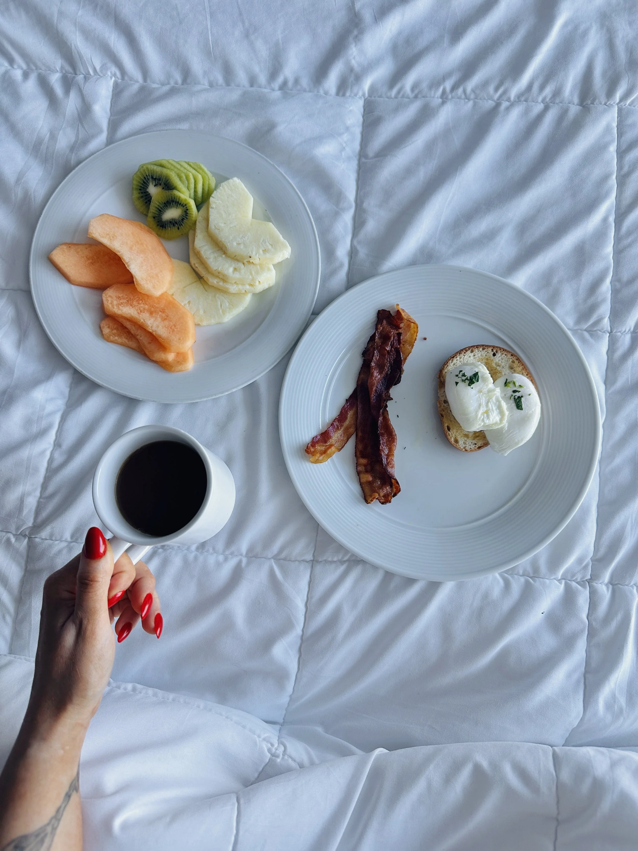 A person with red nail polish holding a cup of black coffee, with breakfast plates on a white quilted bedspread. The plates contain sliced kiwi, melon, apple, a piece of bacon, and an English muffin with poached eggs and herbs.