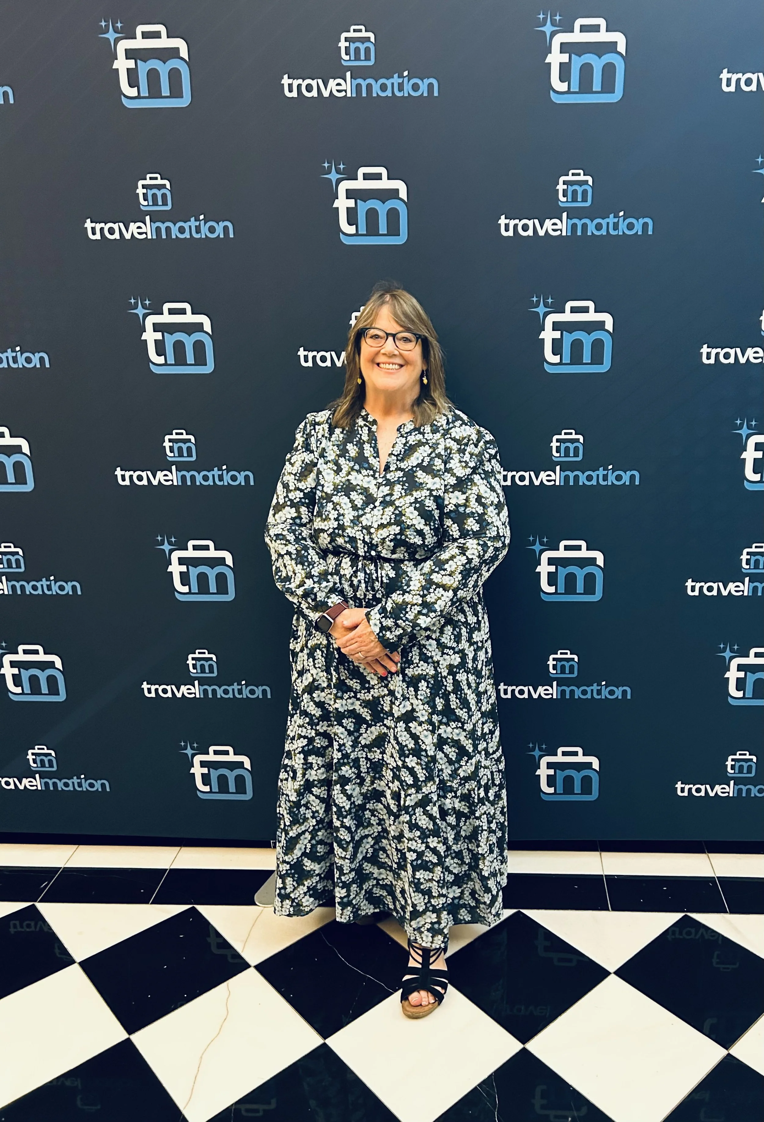 A woman standing in front of a travelmediation branded backdrop, smiling and wearing glasses, a long floral dress, and black sandals, with a black and white checkered floor beneath her.