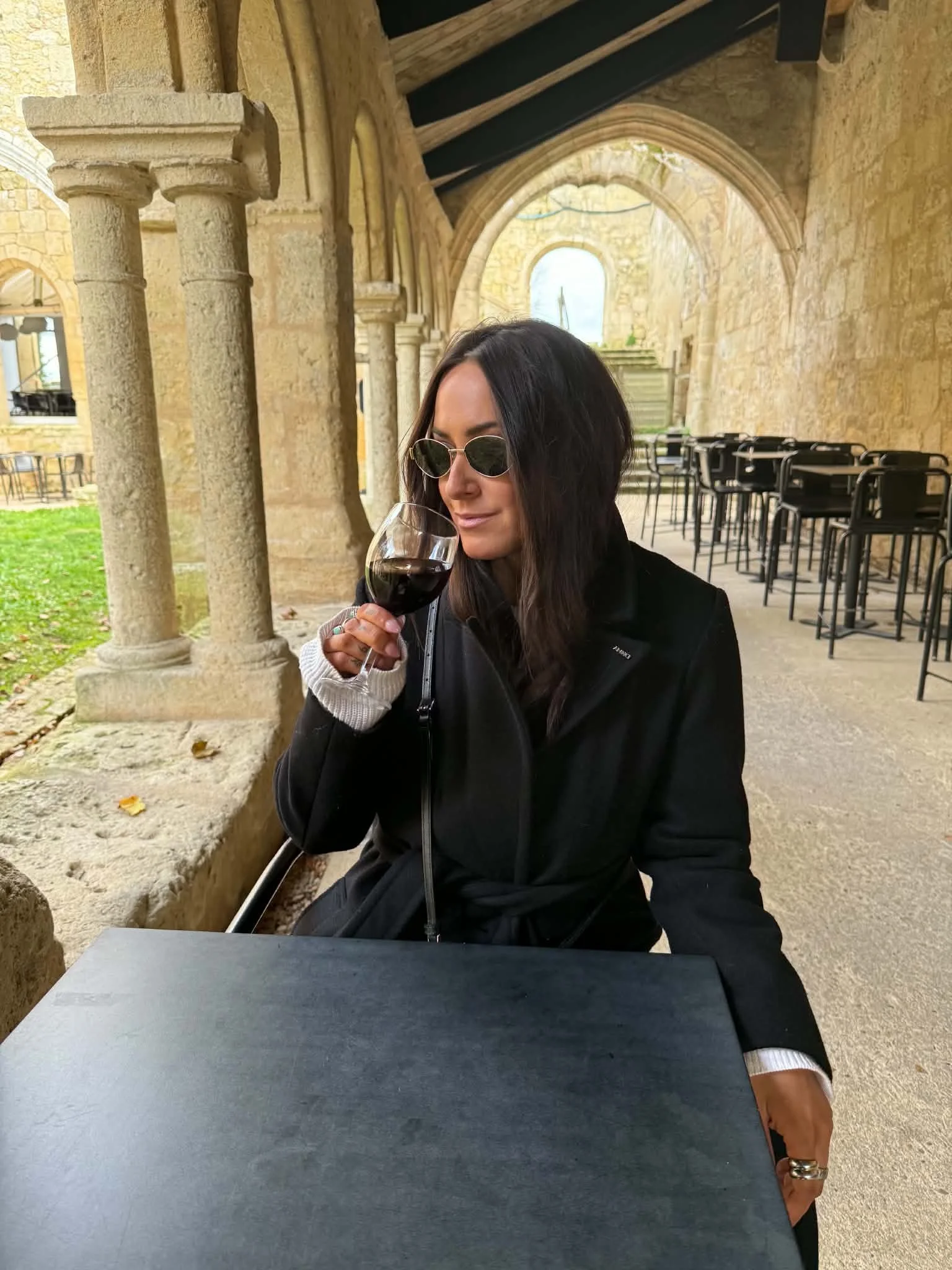 A woman with long dark hair, wearing sunglasses and a black jacket, seated outdoors in a historic stone building with arches, drinking red wine.